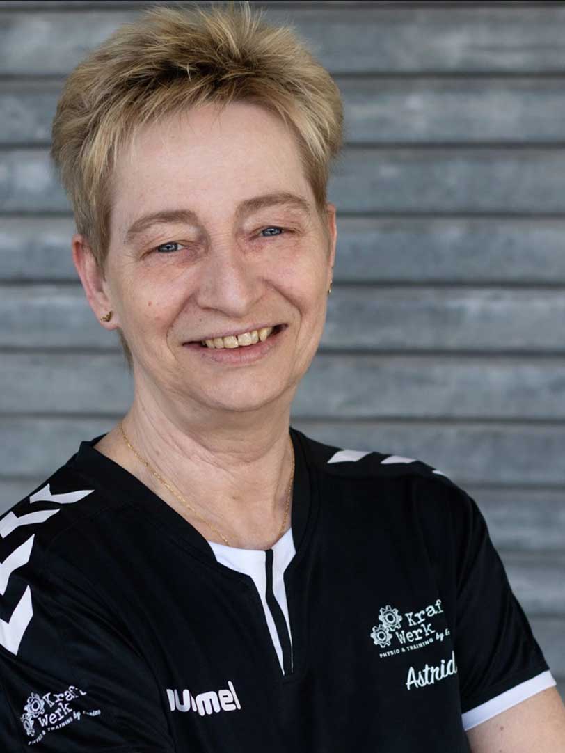 Person mit kurzem blondem Haar lächelt in schwarzem Sportshirt vor grauem Hintergrund