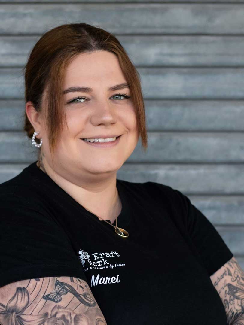 Eine lächelnde Person mit tätowierten Armen trägt ein schwarzes T-Shirt vor einer grauen Wand und hat braune, halblange Haare.